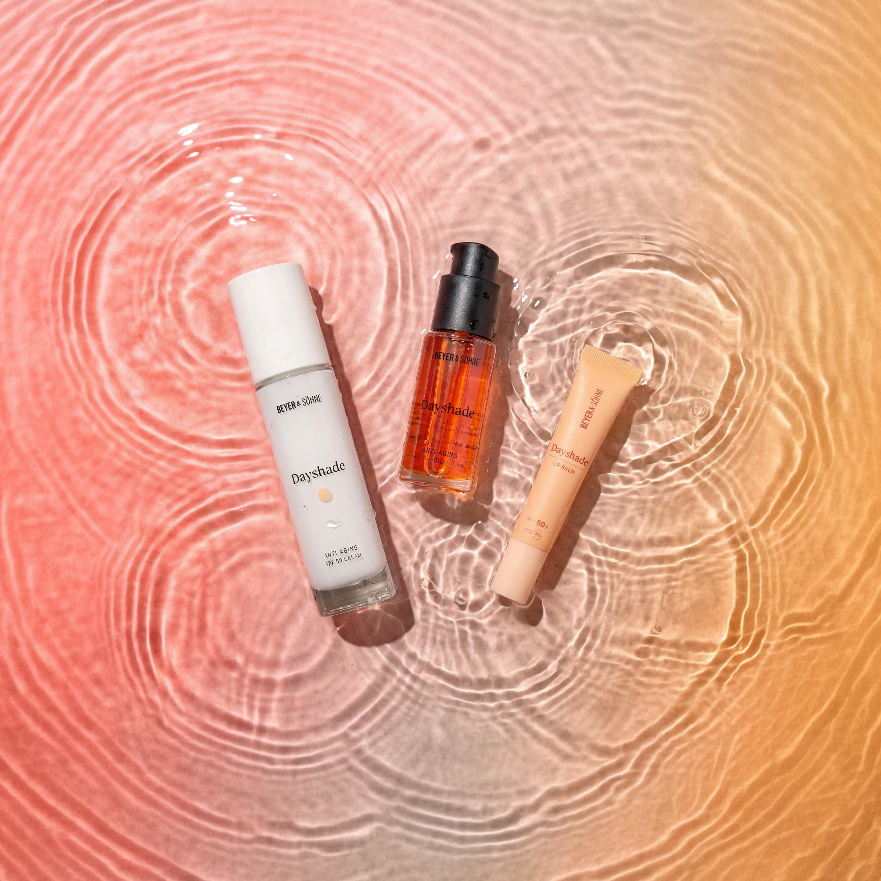 Drei Dayshade Produkte liegen auf einer Wasseroberfläche mit sanften Wellen, umgeben von warmen Farbverläufen in Orange- und Rosétönen, die eine leichte, frühlingshafte Atmosphäre vermitteln.